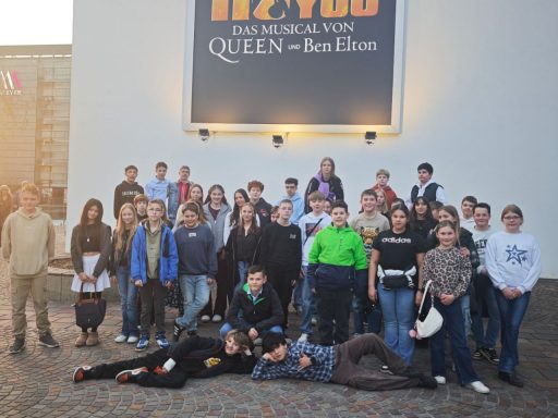 Gruppe von Jugendlichen und Erwachsenen posiert vor einer großen Wand mit Werbung für "Queen".