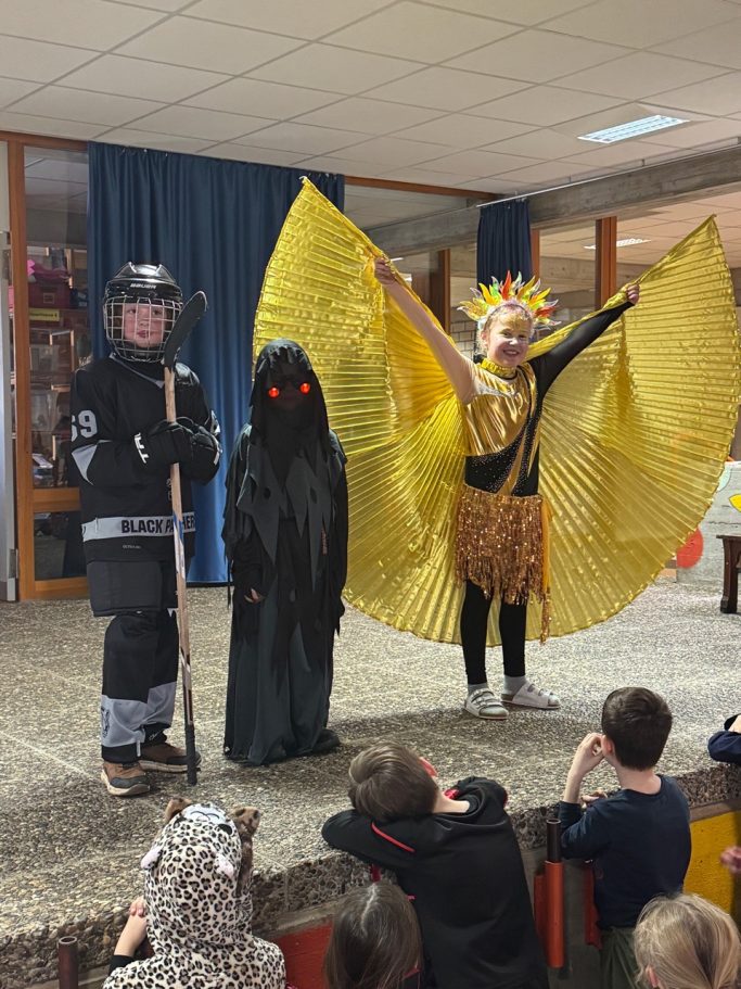 Kostümgewinner in der Grundschule Drei Kinder in Kostümen: Polizei, Gespenst und goldener Vogel mit Flügeln auf einer Bühne.