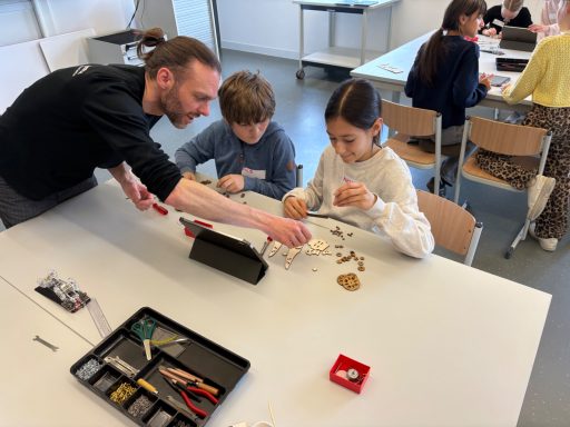 Ein Mitarbeitet zeigt zwei Kinder, wie sie mit Bastelmaterialien arbeiten.