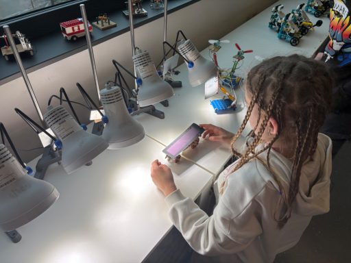 Ein Kind mit Dreadlocks betrachtet ein Smartphone an einem Tisch mit Lampen und Spielzeugmodellen.