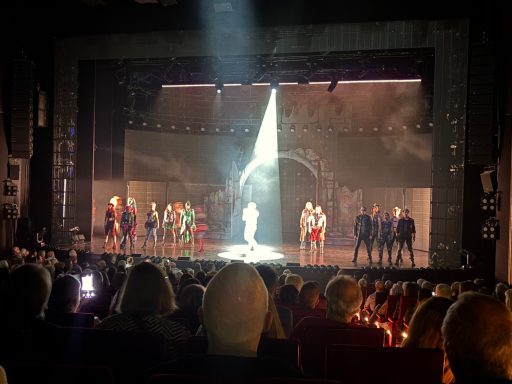 Bühnenauftritt mit Darstellern in bunten Kostümen, beleuchtet von einem Scheinwerfer.