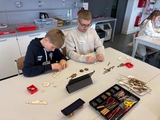 Zwei Kinder basteln mit Materialien auf einem Tisch, umgeben von Werkzeugen und Materialien.